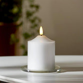 Valódi viaszból készült, fehér színű led gyertya üveg tányéron, fehér színű asztalon, szürke fal előtt